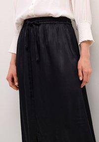 Zwarte satijnen rok met een elastische tailleband en strikdetail. Heeft een zijsplit en een gladde textuur. Gecombineerd met een witte blouse met knopen.