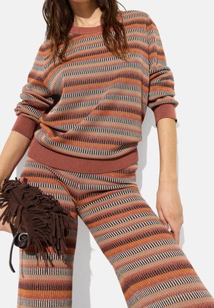 Kobieta ubrana w pasujący sweter i spodnie w paski w kolorach rdzy, pomarańczu, czerni i beżu, trzymająca ciemnobrązową torbę z frędzlami.