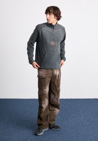 Γκρίζα φλίς ζακέτα με μισό φερμουάρ, μπροστινή τσέπη και πορτοκαλί λογότυπ ο; συνδυασμένη με καφέ cargo παντελόνι που έχει πολλές τσέπες και χαλαρή εφαρμογή.