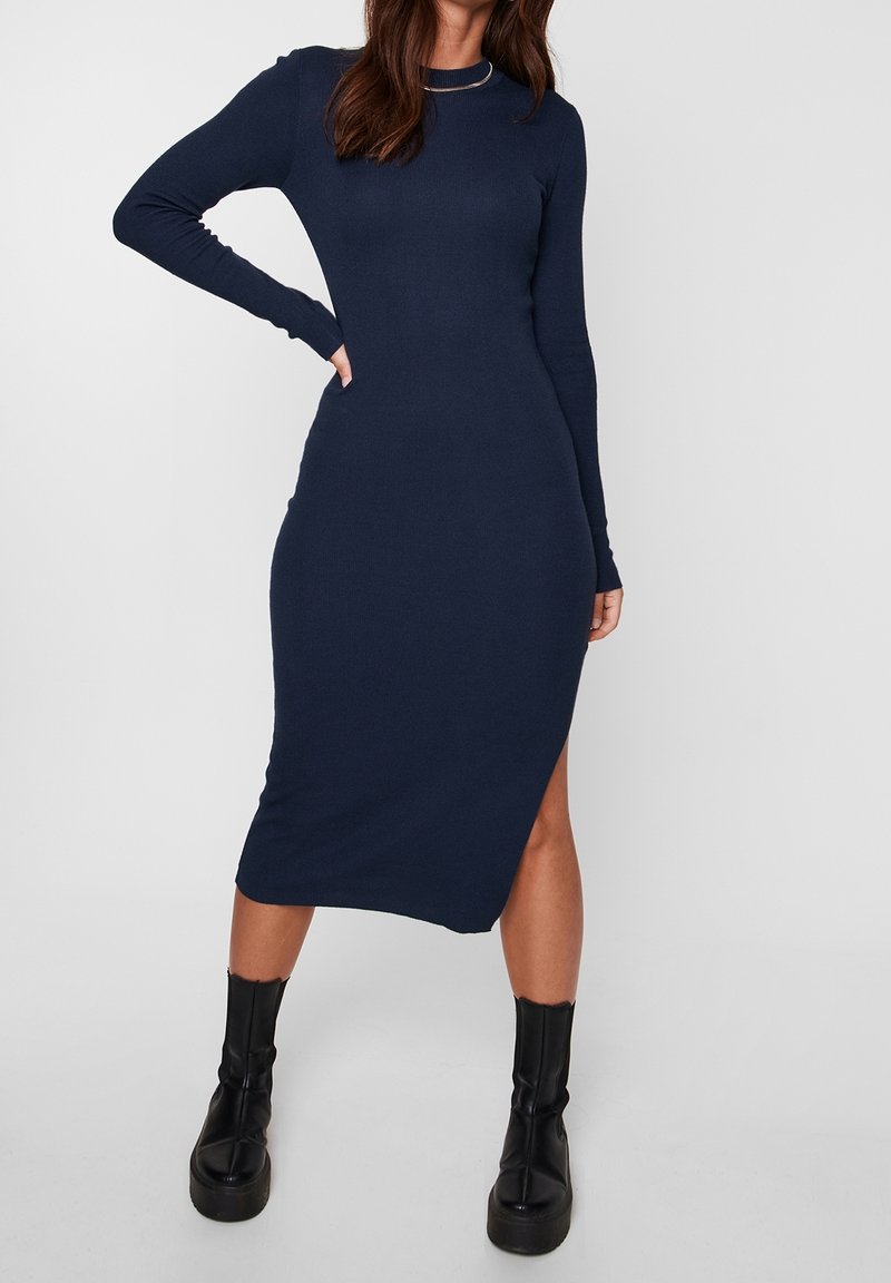 Femme portant une robe midi bleu marine ajustée à manches longues avec une fente latérale et des bottines à plateforme noires, posant avec une main sur la hanche.