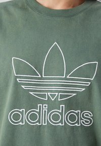 Zielona bawełniana koszulka z białym haftowanym logo Adidas, w tym projekt trzech liści w formie trifolium oraz poziome paski.