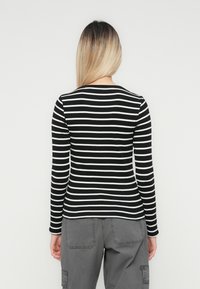 Woman with long blonde hair wearing a black and white striped long-sleeve shirt and gray cargo pants, standing against a plain white background.