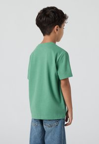 Tricou din bumbac verde, cu mâneci scurte și guler rotund; are o textură netedă și o croială lejeră, asociat cu pantaloni scurți din denim.