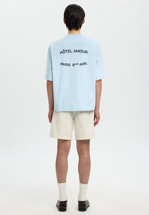 Personne debout de dos portant une chemise bleu clair avec le texte « HÔTEL AMOUR PARIS 9ÈME ARR. », un short beige, des chaussettes blanches et des chaussures foncées.