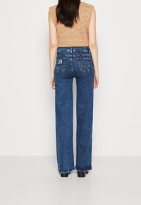 Nainen, joka on pukeutunut korkeavyötäröisiin suoriin sinisiin farkkuihin, joissa on takataskut, ja vaaleanbeigeen ilmanhihaiseen neulepaitaan, seisoo yksiväristä valkoista taustaa vasten.