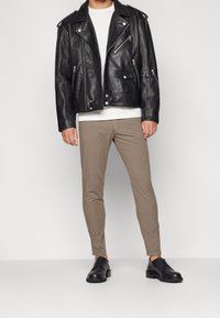 Man som bär en svart läderbikerjacka, vit t-shirt, smala beige byxor och svarta finskor, står mot en neutral bakgrund.