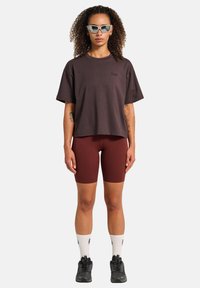 Vrouw met krullend haar die een grijze zonnebril, een losse donkere t-shirt, bordeauxrode fietsshorts, witte sokken en zwarte sneakers draagt, staande met het gezicht naar voren.
