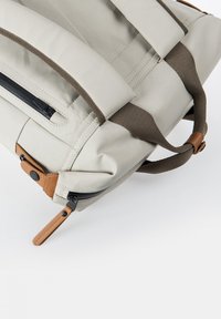 Hellgraue Sporttasche mit dunkelbraunen Griffen, sandfarbenen Lederreißverschluss-Anhängern und schwarzen Reißverschlüssen vor weißem Hintergrund.