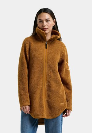 Ung kvinde iført en brun fleecejakke med høj krave og lynlås foran samt blå jeans, stående foran en ensfarvet baggrund.