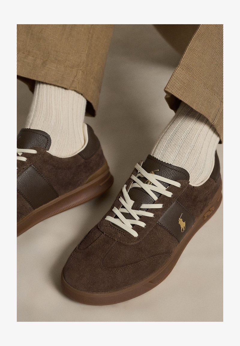 Rudi zomšiniai ir odiniai sportbačiai su baltais raišteliais, dėvimi su smėlio spalvos dryžuotomis kojinėmis ir rudomis tekstūruotomis kelnėmis ant neutralaus fono.