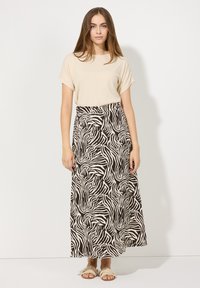 Beige kurzärmliges Oberteil, kombiniert mit einem langen Rock im schwarz-weißen Zebra-Muster. Leichte flache Sandalen. Ein schlichter Hintergrund bietet Kontrast.