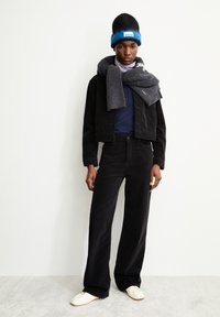 Schwarze Cord-Jacke über einem marineblauen Polohemd, kombiniert mit weiten schwarzen Hosen. Grauer Schal um den Hals gelegt und eine schwarze Mütze mit blauen Akzenten.