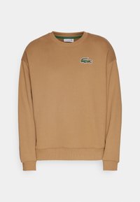 Sudadera de cuello redondo de manga larga de color marrón claro con puños y dobladillo acanalados, que presenta un pequeño logotipo de cocodrilo verde en el pecho izquierdo.