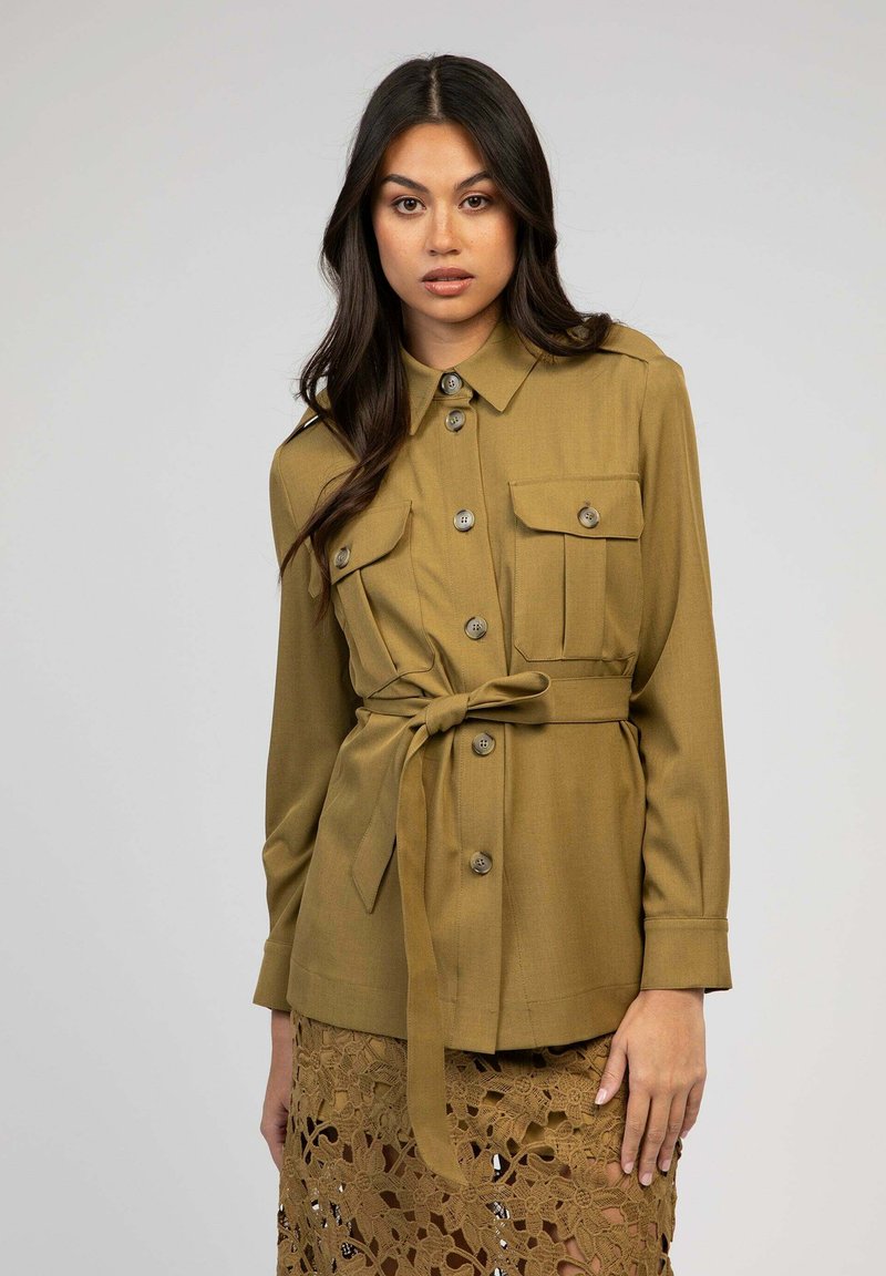 Femme portant une veste en khaki à manches longues et à boutons, avec des poches poitrine et une ceinture nouée à la taille, associée à une jupe en crochet marron.