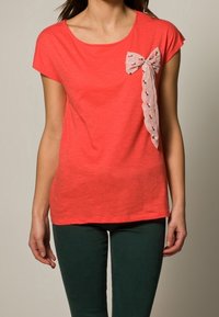 Koralowa bluzka z krótkim rękawem, o okrągłym dekolcie, z wzorzystą, jasnoróżową kokardką po lewej stronie, wykonana z miękkiego, teksturowanego materiału.