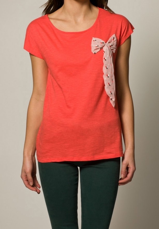 Koralowa bluzka z krótkim rękawem, o okrągłym dekolcie, z wzorzystą, jasnoróżową kokardką po lewej stronie, wykonana z miękkiego, teksturowanego materiału.