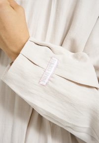Light beige cotton shirt sleeve with a folded cuff, featuring a tag with "BALMOND SPRING" stitched in white and surrounded by pink thread.