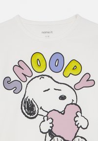 Hvit kortermet t-skjorte med en tegneseriehund som holder et rosa hjerte. Teksten "SNOOPY" i gult, lilla og rosa over figuren.