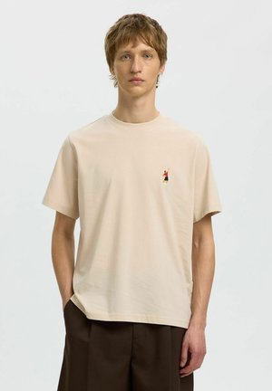 Młody mężczyzna o jasnobrązowych włosach, ubrany w beżowy t-shirt z małą kolorową haftowaną postacią na piersi, stoi na jednolitym tle.