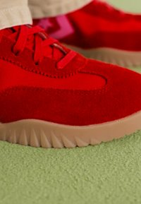 Rød sneakers med en kombination af glat og struktureret materiale, med en gummisål med et mønstret træd og matchende røde snørebånd.