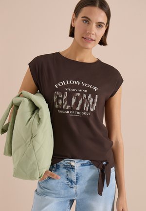 Femme portant un t-shirt noir avec le texte « Follow Your Flow », un jean bleu clair, tenant un sac matelassé vert clair sur l'épaule.