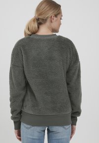 Felpa grigia in pile con una superficie texturizzata e soffice. Presenta polsini e orlo a coste, e una vestibilità casual. Indossata sopra jeans azzurri chiari.