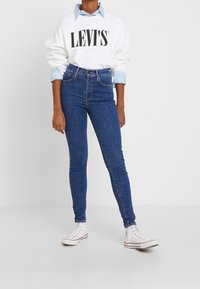 Sudadera blanca con el logo negro de "LEVI'S", superpuesta sobre una camisa de color azul claro; combinada con jeans oscuros ajustados y zapatillas blancas.