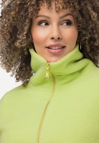 Limonengrüne Fleecejacke mit hohem Kragen und goldener Reißverschluss. Weiche Textur und minimalistisches Design.