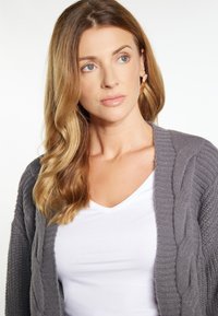 Cardigan grigio in maglia con motivo a treccia, caratterizzato da una finitura texture. Indossato sopra una t-shirt bianca a scollo a V. Fibra morbida con un taglio rilassato.