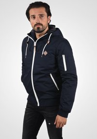 Marineblauwe jas van stof met een ritssluiting, witte accenten en meerdere zakken; voorzien van een capuchon en geborduurd embleem op de borst.