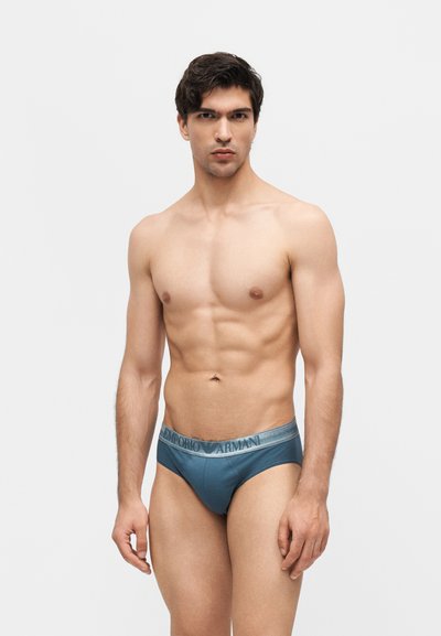 Άνδρας χωρίς πουκάμισο με κοντά σκούρα μαλλιά που φοράει μπλε εσώρουχα Emporio Armani, στέκεται απέναντι σε ένα απλό λευκό φόντο.