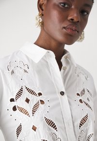 Camicia bianca con dettagli di ricamo intagliato intricati. Presenta un colletto classico e bottoni decorativi dorati.