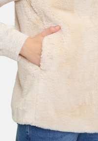 Cremfarbene Kunstfelloberteil mit weicher, flauschiger Textur und einer Seitentasche. Rippbündchen und lockere Passform, ideal für Freizeitbekleidung.