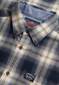Chemise à carreaux bleu et beige à boutons marron, avec col plié et écusson Superdry Co. Machined sur la poche poitrine.