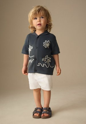 Toddler with blonde hair wears a dark short-sleeve buttoned shirt with sun and palm tree designs, white shorts, and dark sandals, standing.