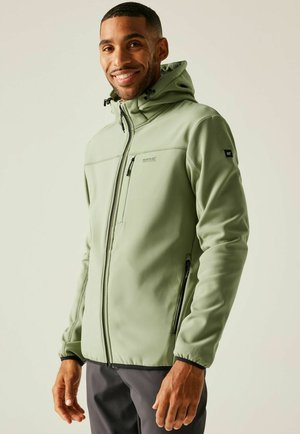 Hombre sonriente con chaqueta al aire libre con capucha y cremallera de color verde claro con bolsillos con cremallera y pantalones gris oscuro, de pie contra un fondo claro.