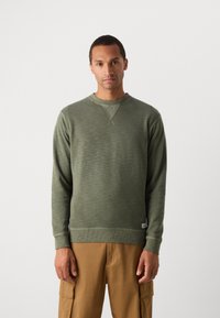 Olijfgroene gebreide sweatshirt met een ronde hals, ribgebreide mouwen en onderkant. Heeft een subtiele textuur en een label detail aan de onderlinkerkant.