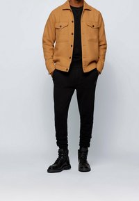 Brun ullblandad jacka med två bröstfickor, svart tröja under, svarta sweatpants med mudd, och svarta kängor.