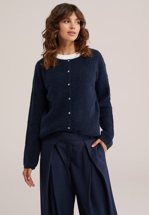 Frau mit welligem braunem Haar, die eine marineblaue flauschige, geknöpfte Strickjacke über einem weißen Top und dunklen plissierten weiten Hose trägt, Hand in der Tasche, schaut nach rechts.