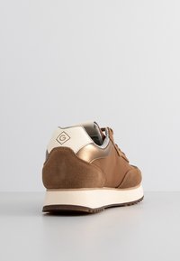 Une basket marron unique avec des panneaux en daim et métal, une semelle blanche et un languette de talon beige affichant un logo diamant, vue de dos sur fond uni.