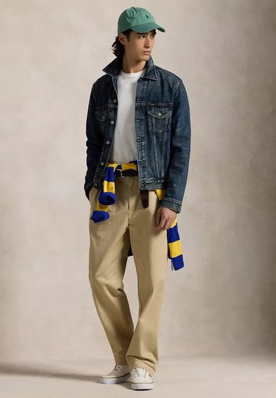 Giacca di jeans, t-shirt bianca, pantaloni khaki e maglione a righe blu-gialle annodato in vita; accessoriato con un cappellino teal e sneakers bianche.