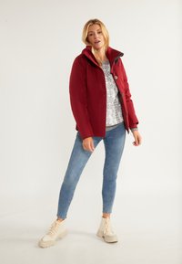 Rød isoleret jakke med lynlås foran, grå sweater med mønsterdesign, blå skinny jeans og lyse snørebåndsstøvler.