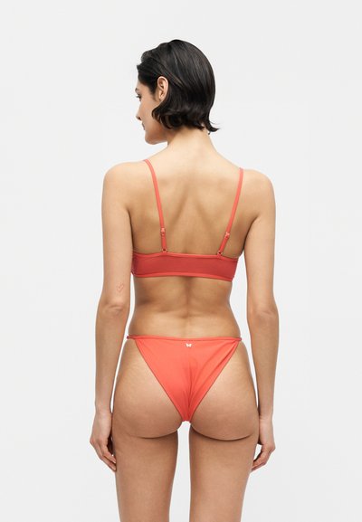 Γυναίκα με κοντά σκούρα μαλλιά, που φοράει κόκκινο μπικίνι με λεπτές τιράντες, στέκεται με την πλάτη προς ένα απλό ανοιχτό γκρι φόντο.