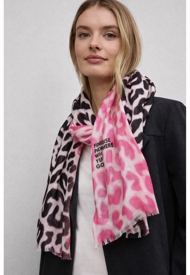 Femme portant un manteau noir et une écharpe à imprimé léopard rose et noir avec le texte "FIND RISE PART HERE WHERE YOU GO" sur un fond uni.