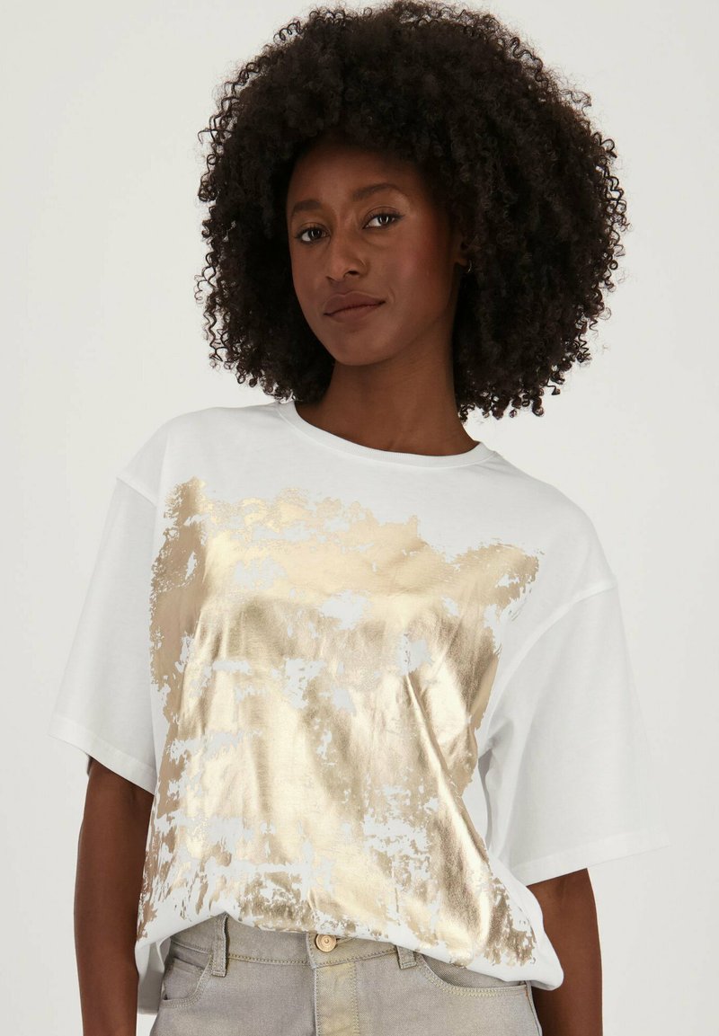 Witte T-shirt met korte mouwen featuring een grote gouden metallic grafiek. Ontspannen pasvorm met een ronde halslijn en een zachte stofstructuur.