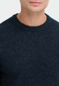 Tume roheline soonikkoes, millel on ümar kaelus ja tekstuuriga kangas, rõhutades pehmet ja sooja välimust.