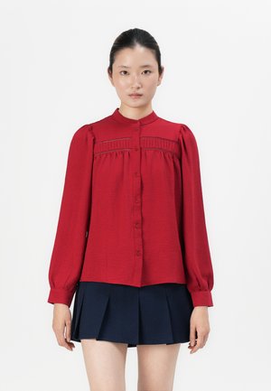 Woman standing, wearing a red long-sleeve buttoned blouse with lace details and a navy blue pleated skirt against a white background.