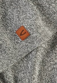 Gray knitted fabric with a textured pattern, featuring a square brown leather patch embossed with a logo.