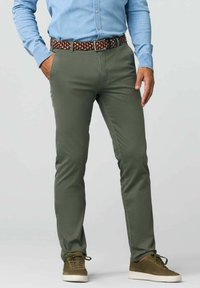 Olivgrüne elastische Hosen mit schmaler Passform, kombiniert mit einem gemusterten gewebten Gürtel. Das Model trägt braune Sneakers mit hellen Sohlen.