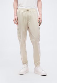 Μπεζ ελαφριά joggers με ελαστική μέση, κορδόνι για ρύθμιση, πλαϊνές τσέπες cargo και στενή εφαρμογή. Συνδυασμένα με λευκά αθλητικά παπούτσια.